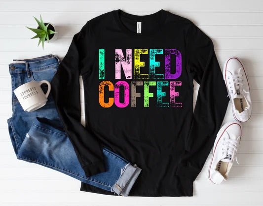 Flat lay of a black long-sleeve shirt with colorful bold text reading “I NEED COFFEE,” styled with jeans and sneakers.